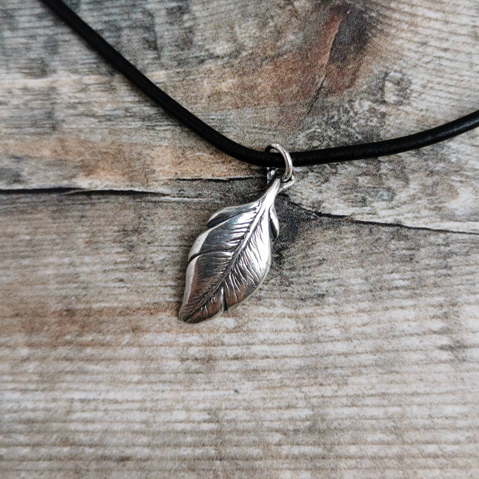 A silver feather pendant necklace on a black leather cord, displayed on a rustic weathered wood background.