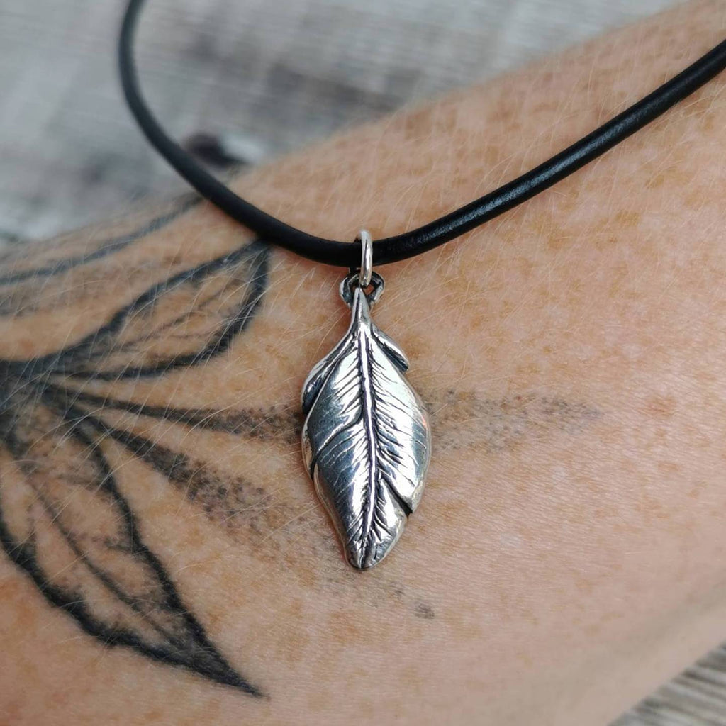 A silver feather pendant necklace on a black leather cord, shown worn on a person's tattooed arm.
