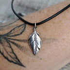 A silver feather pendant necklace on a black leather cord, shown worn on a person's tattooed arm.