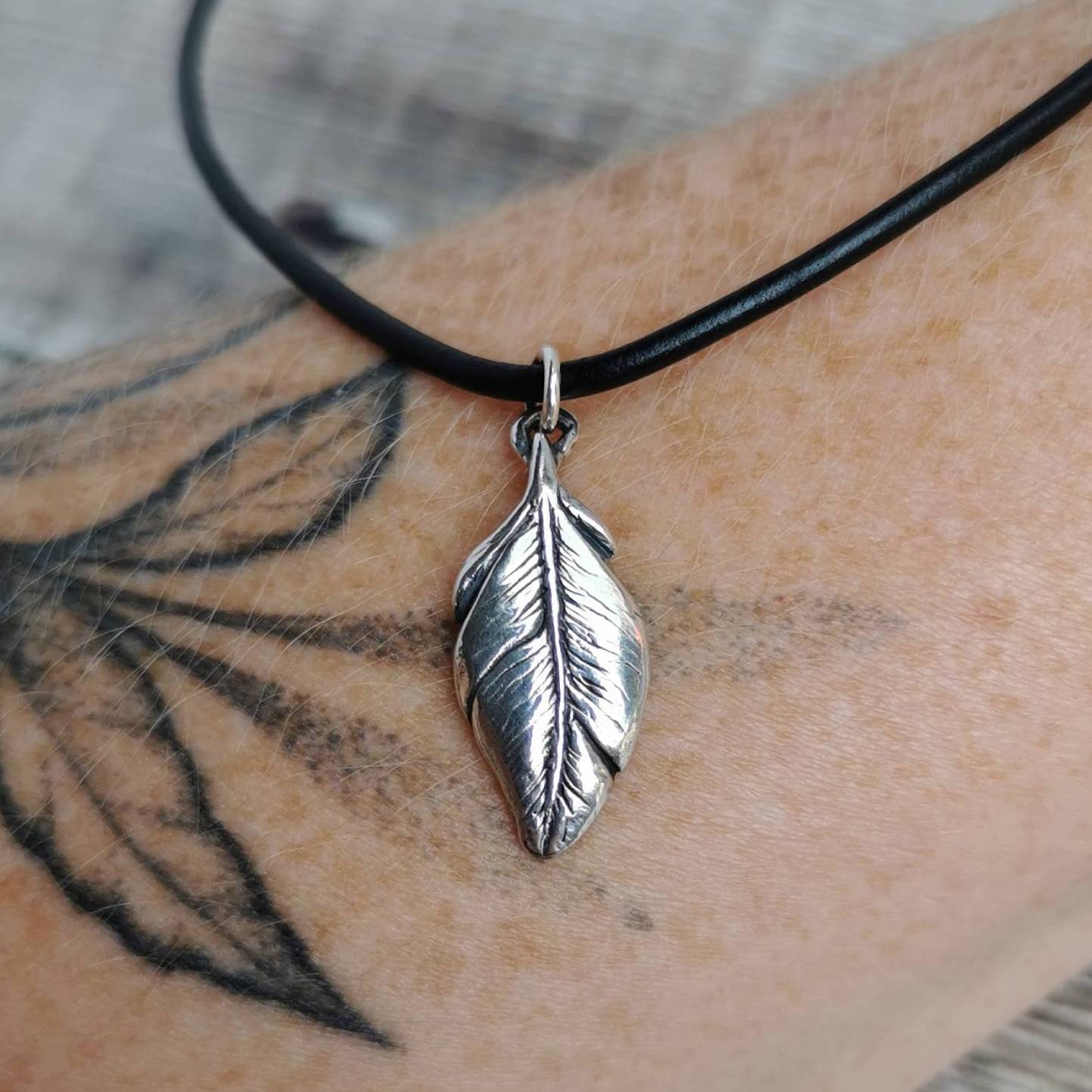 A silver feather pendant necklace on a black leather cord, shown worn on a person's tattooed arm.