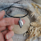 A hand holding a silver feather pendant on a thin black leather cord necklace, with dried grass and a rustic wooden surface in the background.