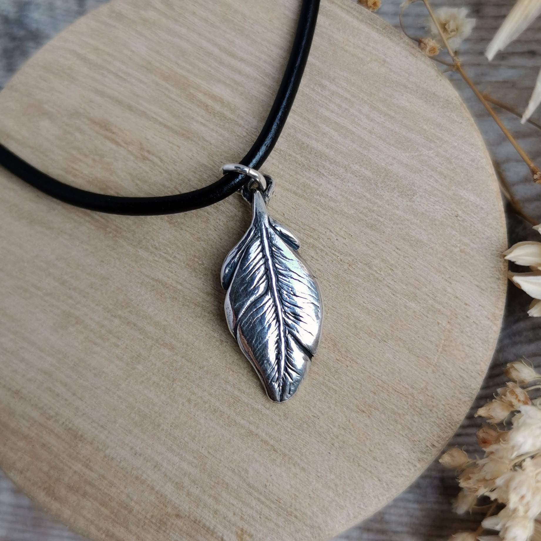 A men's silver feather pendant necklace on a black leather cord, displayed on a round wooden block with dried flowers on a rustic wooden surface.