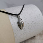 A silver feather pendant necklace on a black leather cord, displayed on a speckled white and beige cylindrical stand.