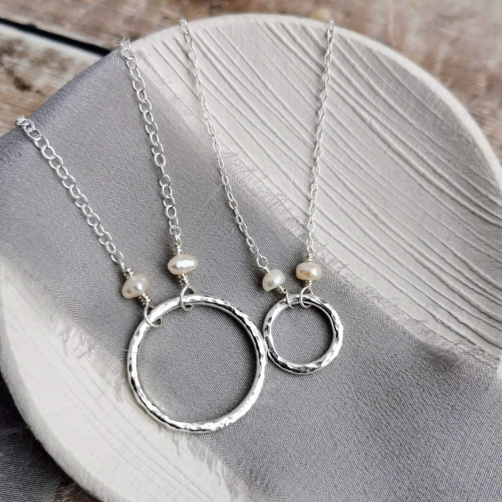 large and small Silver and pearl necklaces with hammered circular pendants and pearls on a gray fabric background