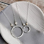 large and small Silver and pearl necklaces with hammered circular pendants and pearls on a gray fabric background
