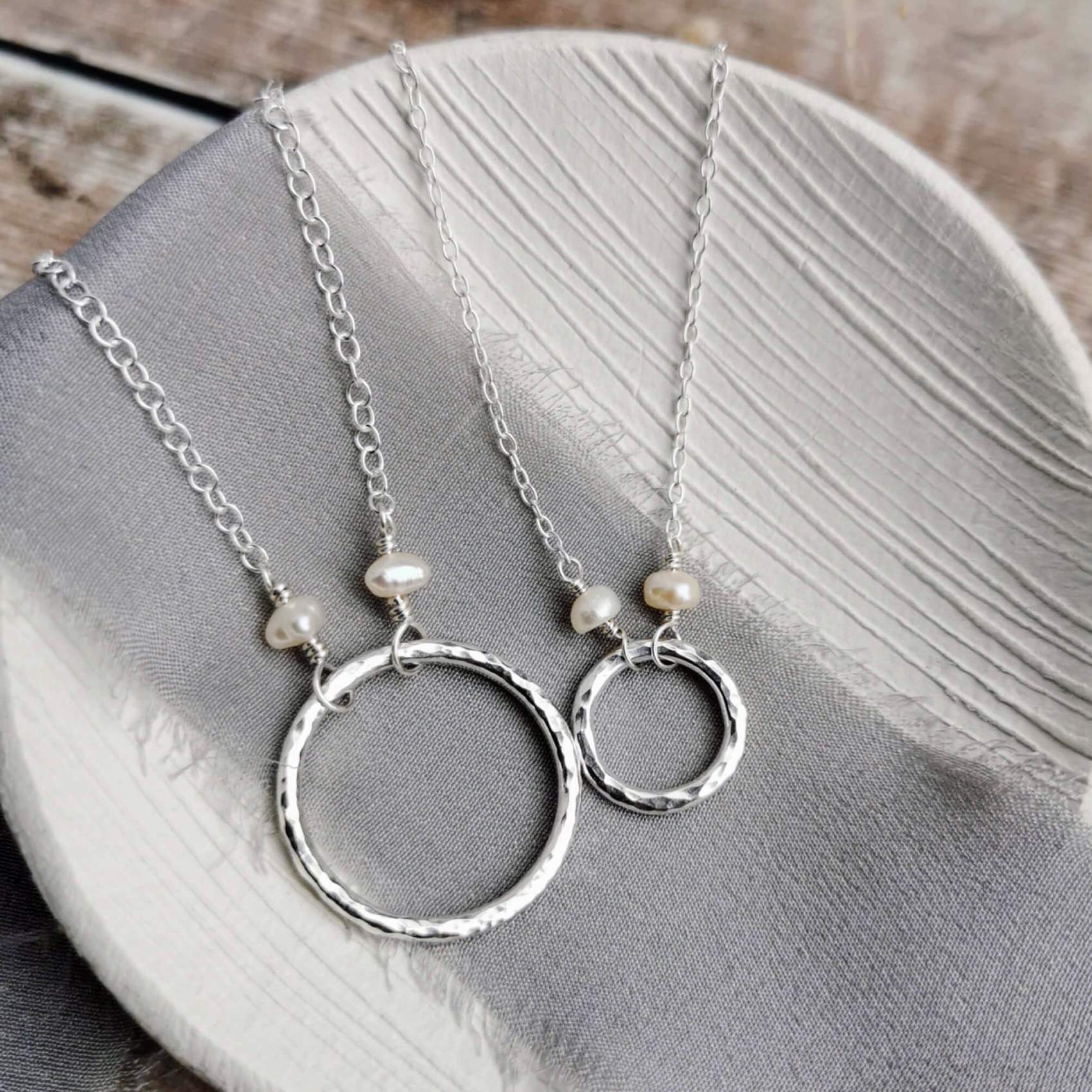 large and small Silver and pearl necklaces with hammered circular pendants and pearls on a gray fabric background