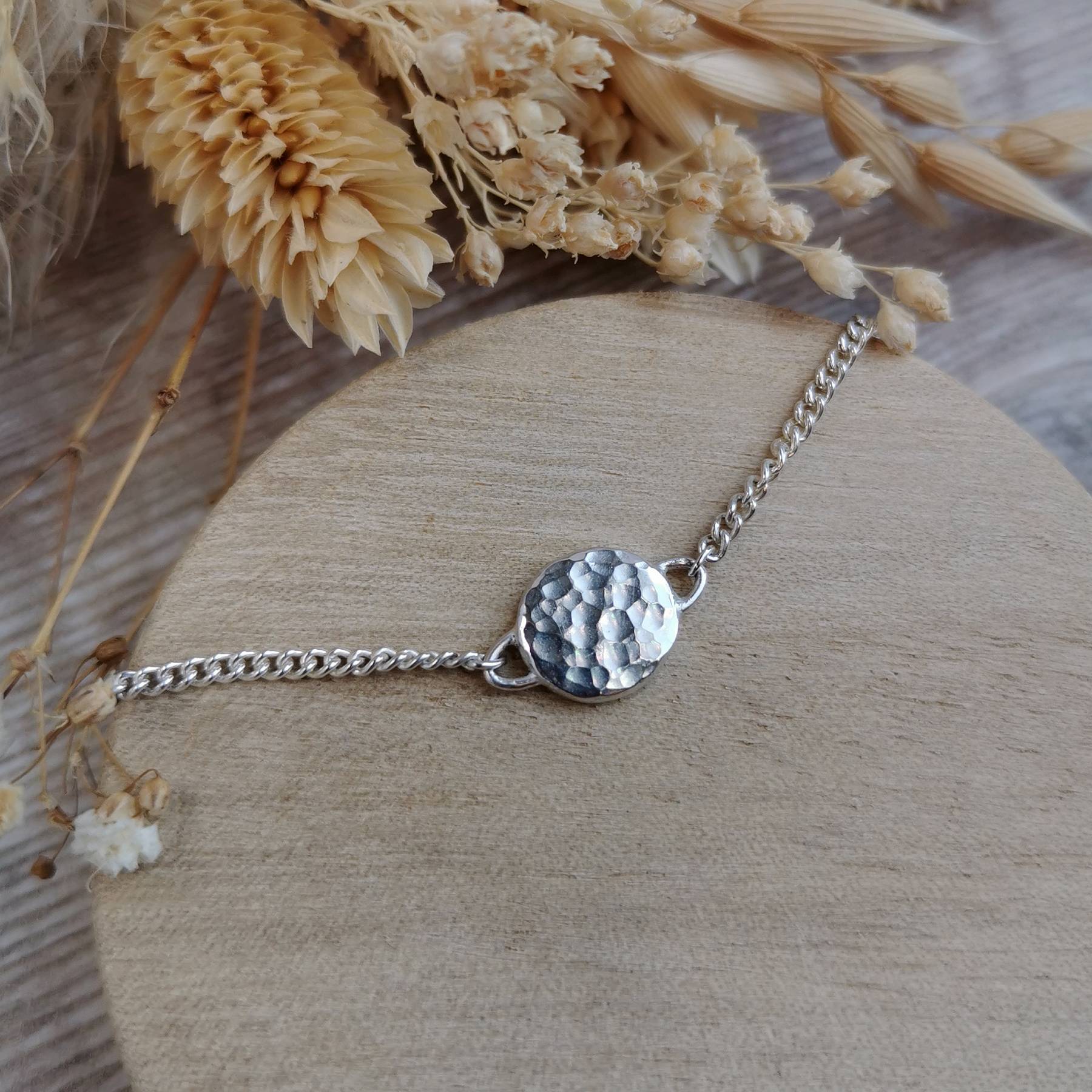 A silver chain bracelet with a textured, hammered circular charm, with a secret word of affirmation hidden on the other side out of sight.  The bracelet is displayed on a round wooden block with dried flowers in the background.