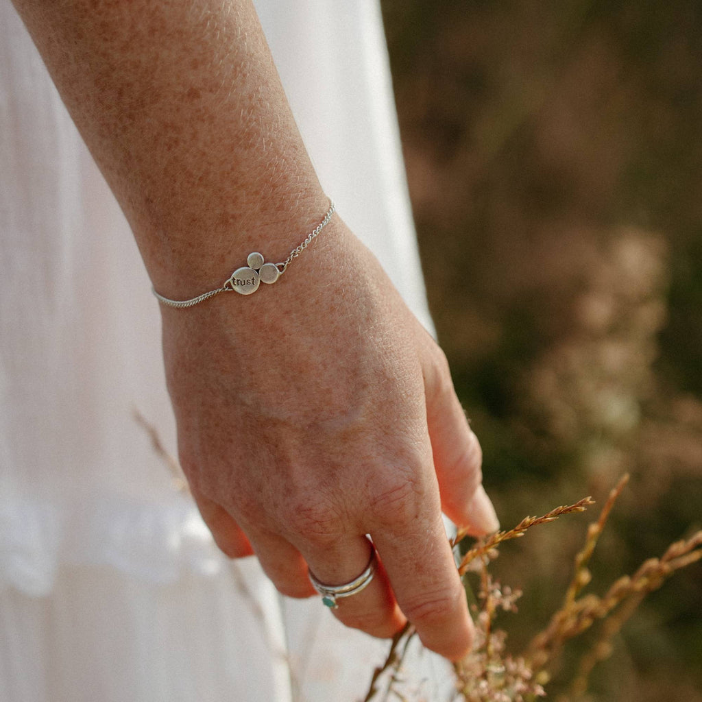 A close-up of a person's hand wearing a delicate silver chain bracelet with three small silver flattened circles, one of which is engraved with the word 'trust'. The person is wearing a white dress and silver and gemstone stacking rings, gently touching dry grass in a field in Cornwall, England.