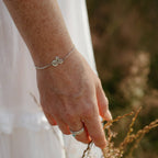 A close-up of a person's hand wearing a delicate silver chain bracelet with three small silver flattened circles, one of which is engraved with the word 'trust'. The person is wearing a white dress and silver and gemstone stacking rings, gently touching dry grass in a field in Cornwall, England.