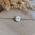 A silver chain bracelet with a round stamped metal disc charm spelling 'align', displayed on a light wood surface with dried flowers in the background.