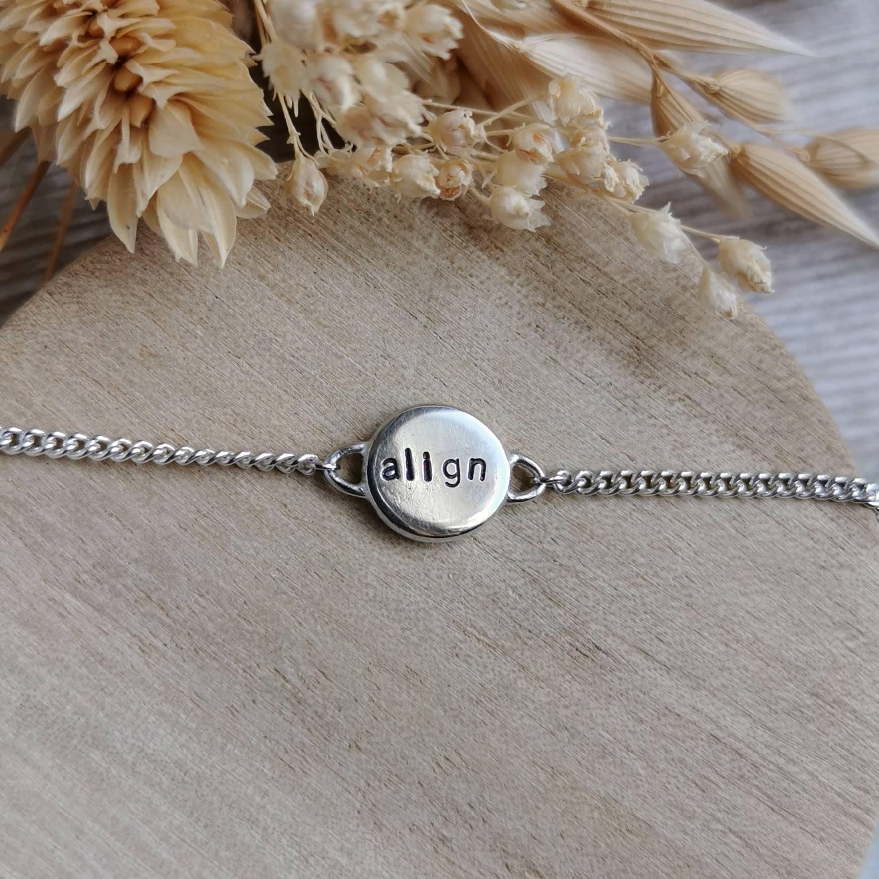 A silver chain bracelet with a round stamped metal disc charm spelling 'align', displayed on a light wood surface with dried flowers in the background.