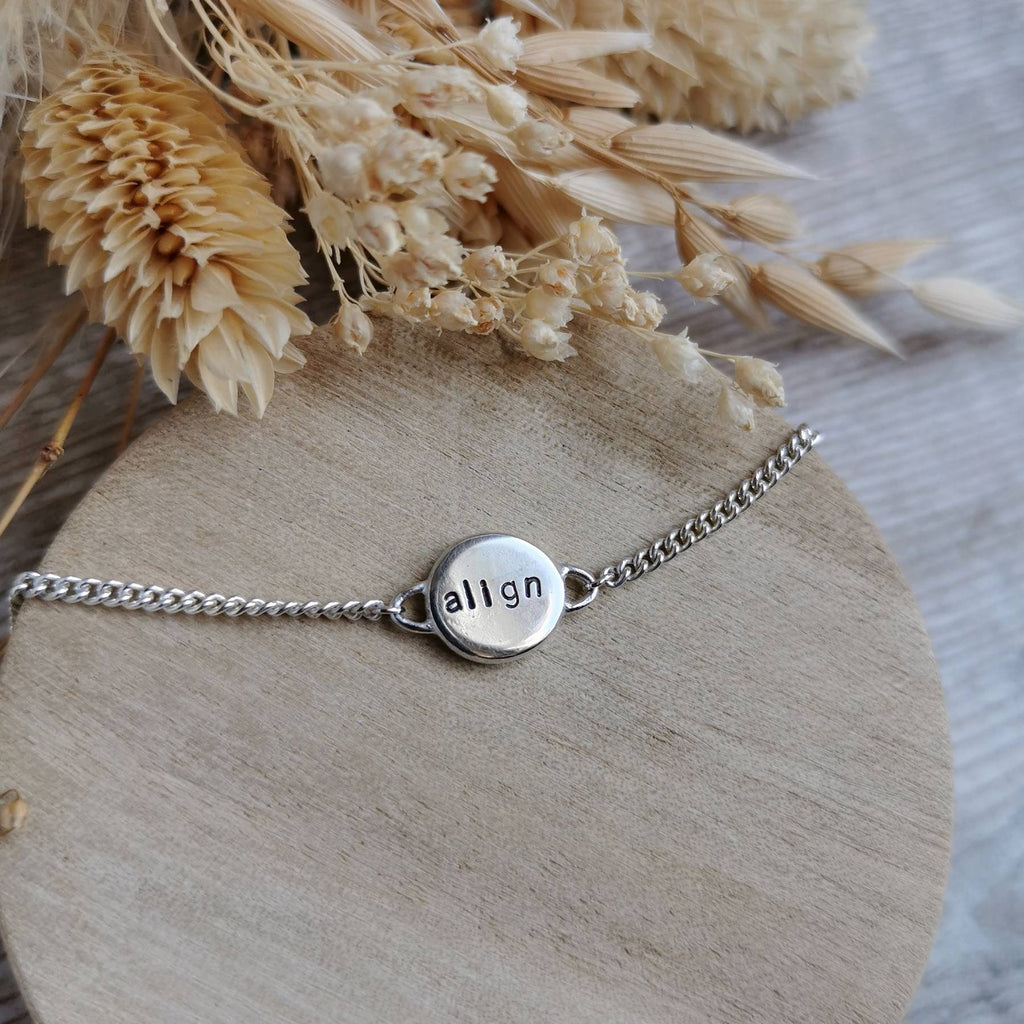 A silver chain mini mantra bracelet with a round engraved disc charm spelling the affirmation 'align', displayed on a round wooden stand with dried flowers in the background.