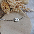 A silver chain mini mantra bracelet with a round engraved disc charm spelling the affirmation 'align', displayed on a round wooden stand with dried flowers in the background.