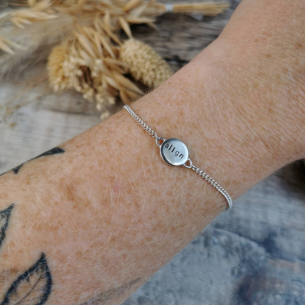 A close-up of a delicate silver chain bracelet worn on a person's wrist, featuring a small round silver disc charm stamped with the affirmation 'align'. The background includes a rustic wooden surface and dried grass.