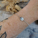 A close-up of a delicate silver chain bracelet worn on a person's wrist, featuring a small round silver disc charm stamped with the affirmation 'align'. The background includes a rustic wooden surface and dried grass.