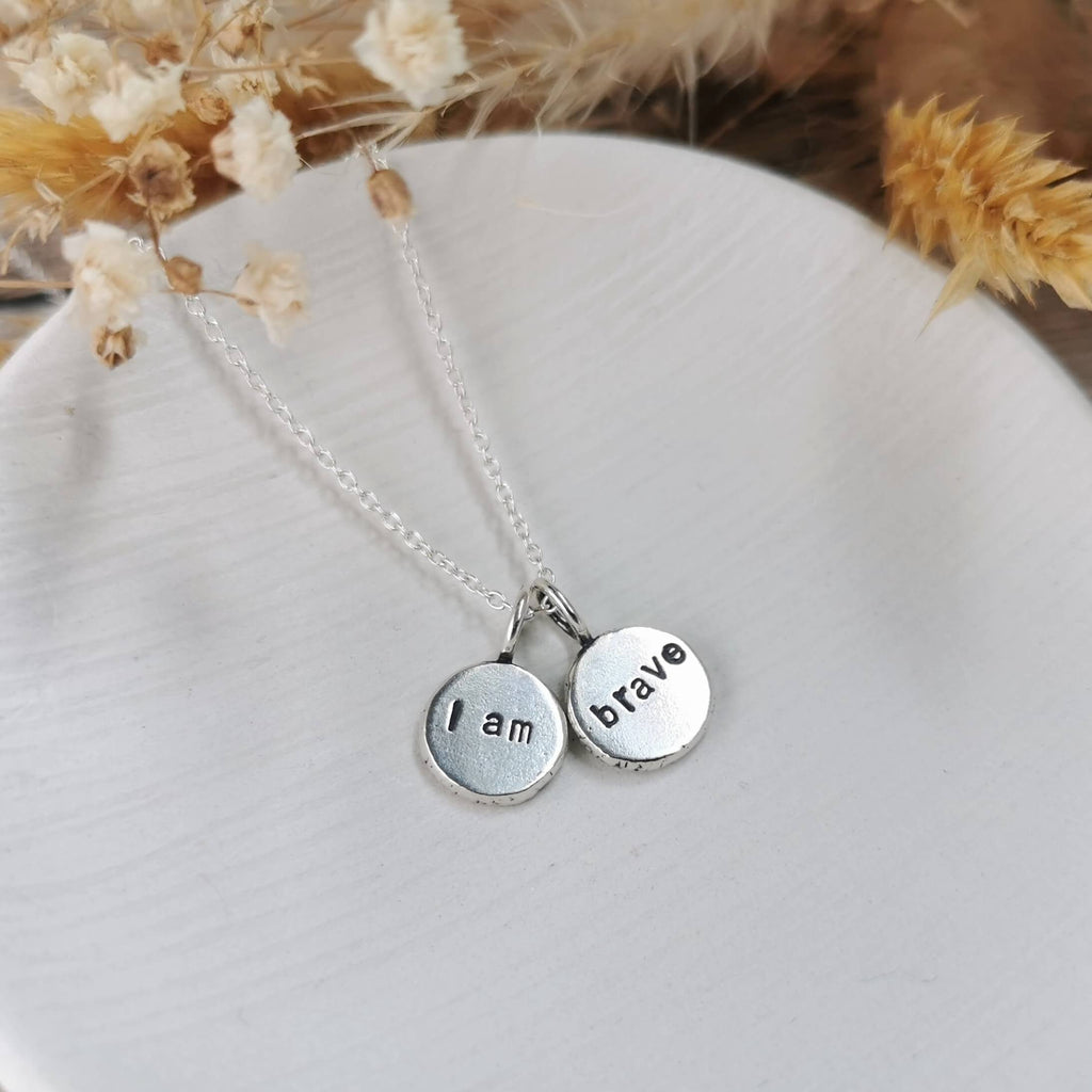 Silver necklace with 'I am brave' affirmation pendants on a white surface with dried flowers in the background