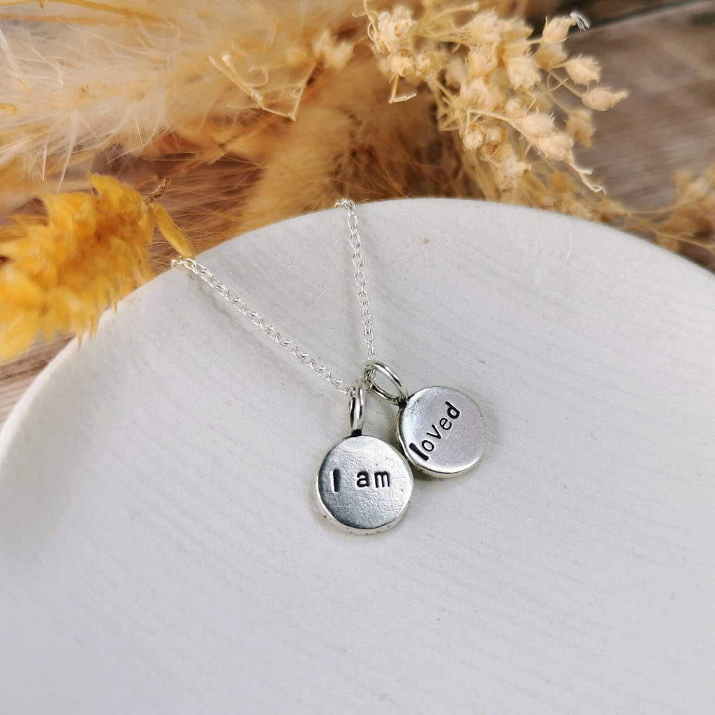 Silver necklace with two round affirmations pendants with the words 'i am' 'loved' on a white surface with dried flowers in the background
