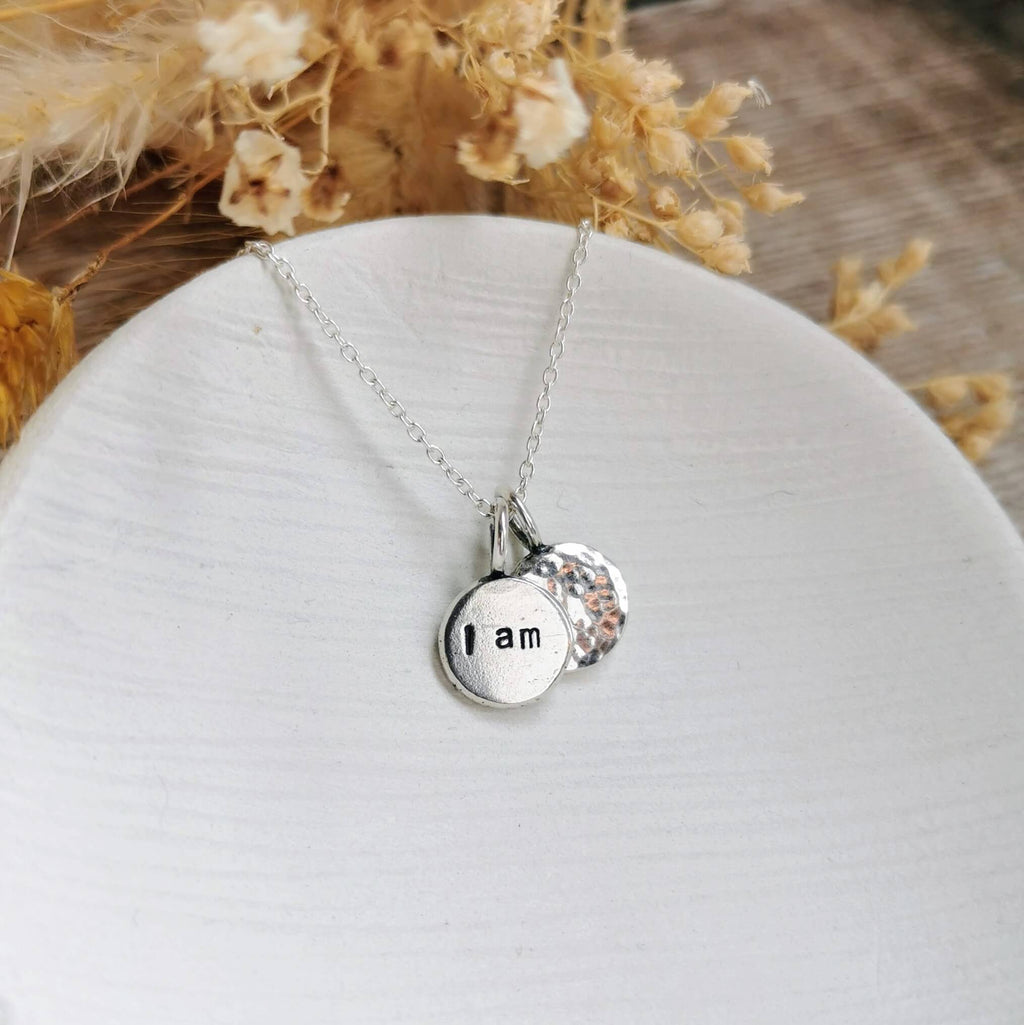 Silver necklace with a round pendant with the words 'I am' with another hammered charm behind, on a white surface with dried flowers in the background