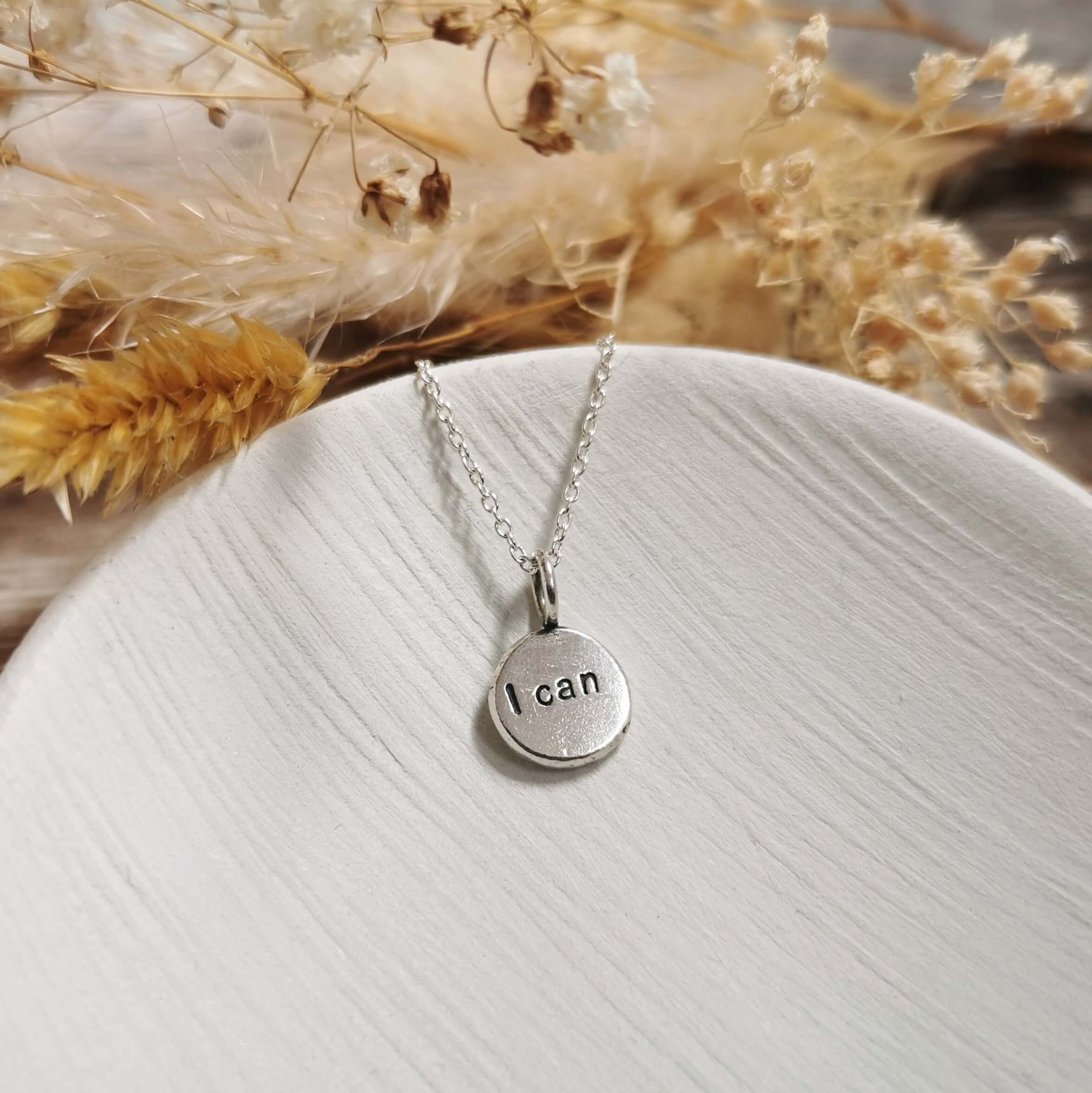 Silver necklace with 'I can' pendant on a white surface with dried flowers in the background