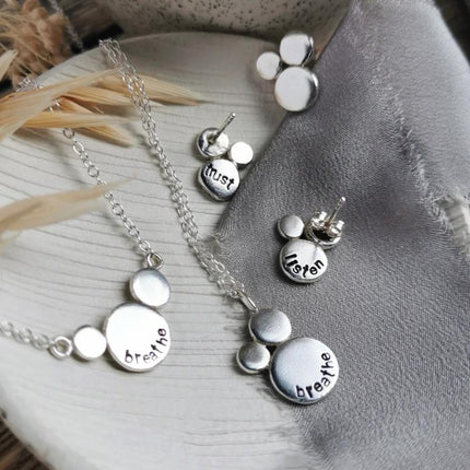 Silver jewelry mini mantra set with necklaces and earrings, all with words of affirmations stamped on them, laid on a textured surface
