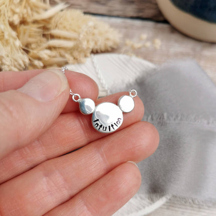 Silver necklace stamped with a custom mini mantra 'Intuition' held in a hand, with blurred natural background