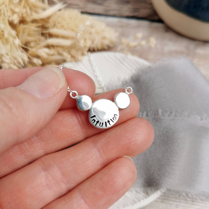Silver necklace stamped with a custom mini mantra 'Intuition' held in a hand, with blurred natural background