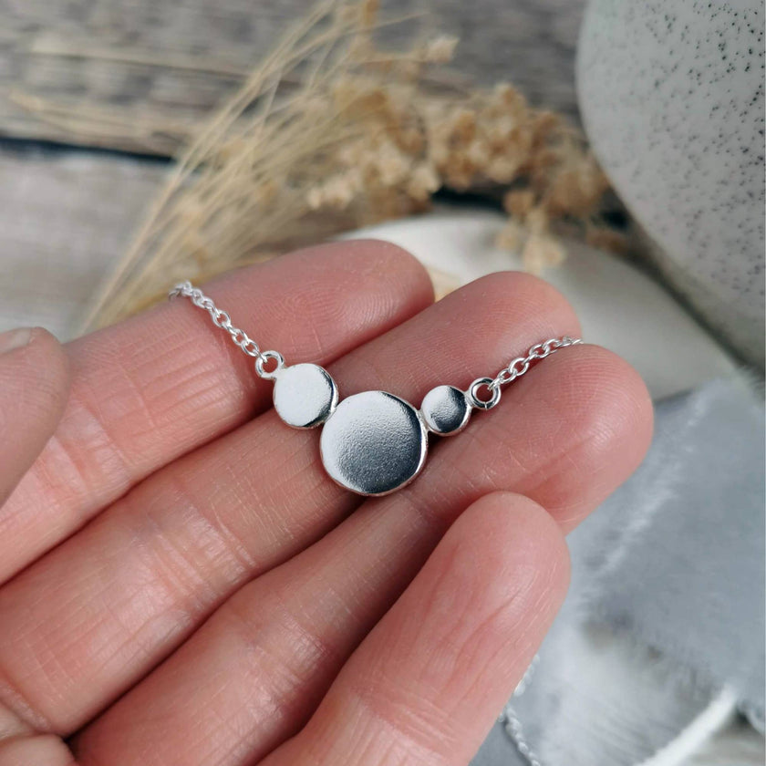 Silver necklace made of circles of recycled silver held in fingers against a blurred natural background