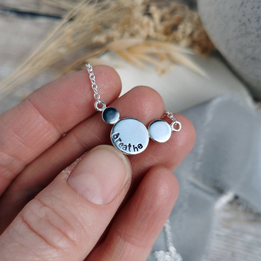 Silver necklace with round silver circles with the word Breathe stamped on, held over a blurred natural background