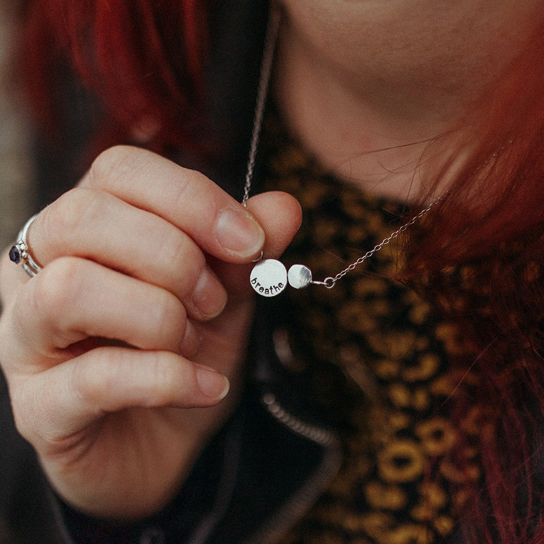 red headed woman wearing silver rings holding a handmade silver necklace with the word Breathe stamped into it against a blurred background