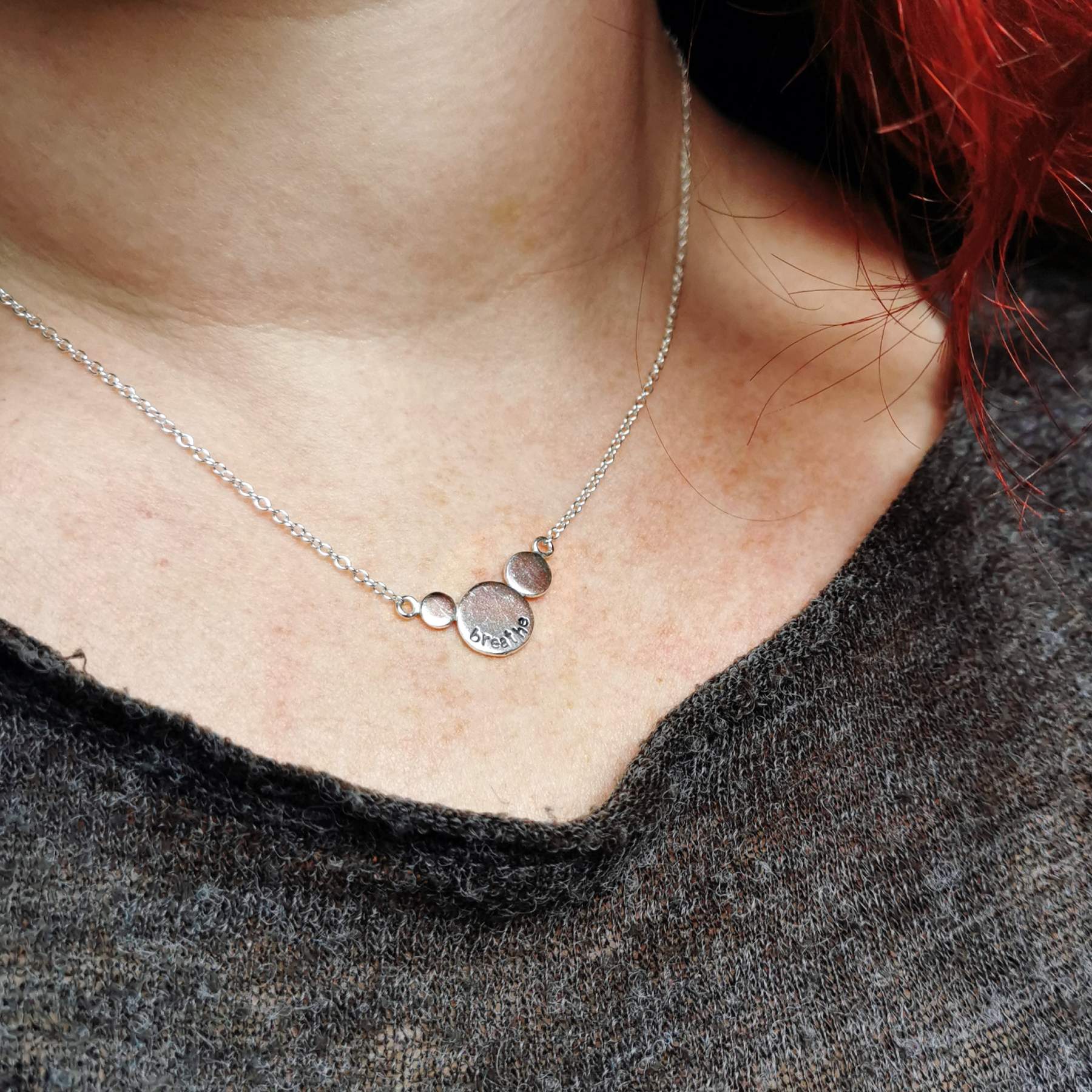Silver necklace with round circles with the word breathe stamped on, worn by a person wearing a dark grey top.