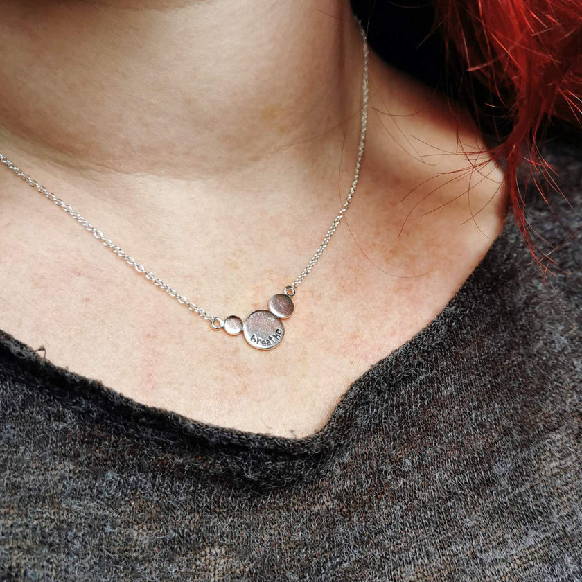 Silver necklace with round circles with the word breathe stamped on, worn by a person wearing a dark grey top.