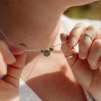A close-up lifestyle shot of a person wearing a delicate sterling silver chain necklace with a central cluster of silver circles in varying sizes, with the largest central one with the word 'breathe' hand stamped onto it. The necklace is being held up by two hands, with silver midi and thumb rings visible on one hand.