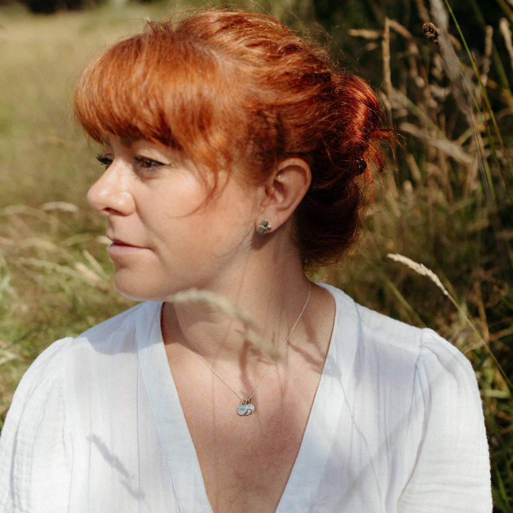 A woman with red hair tied back in a bun, wearing a white blouse and a small silver necklace with a cluster of silver charms, photographed outdoors in a grassy field.