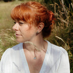 A woman with red hair tied back in a bun, wearing a white blouse and a small silver necklace with a cluster of silver charms, photographed outdoors in a grassy field.