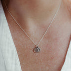 A close-up of a person's neck wearing a delicate silver chain necklace with a small round engraved pendant with the words 'I am'