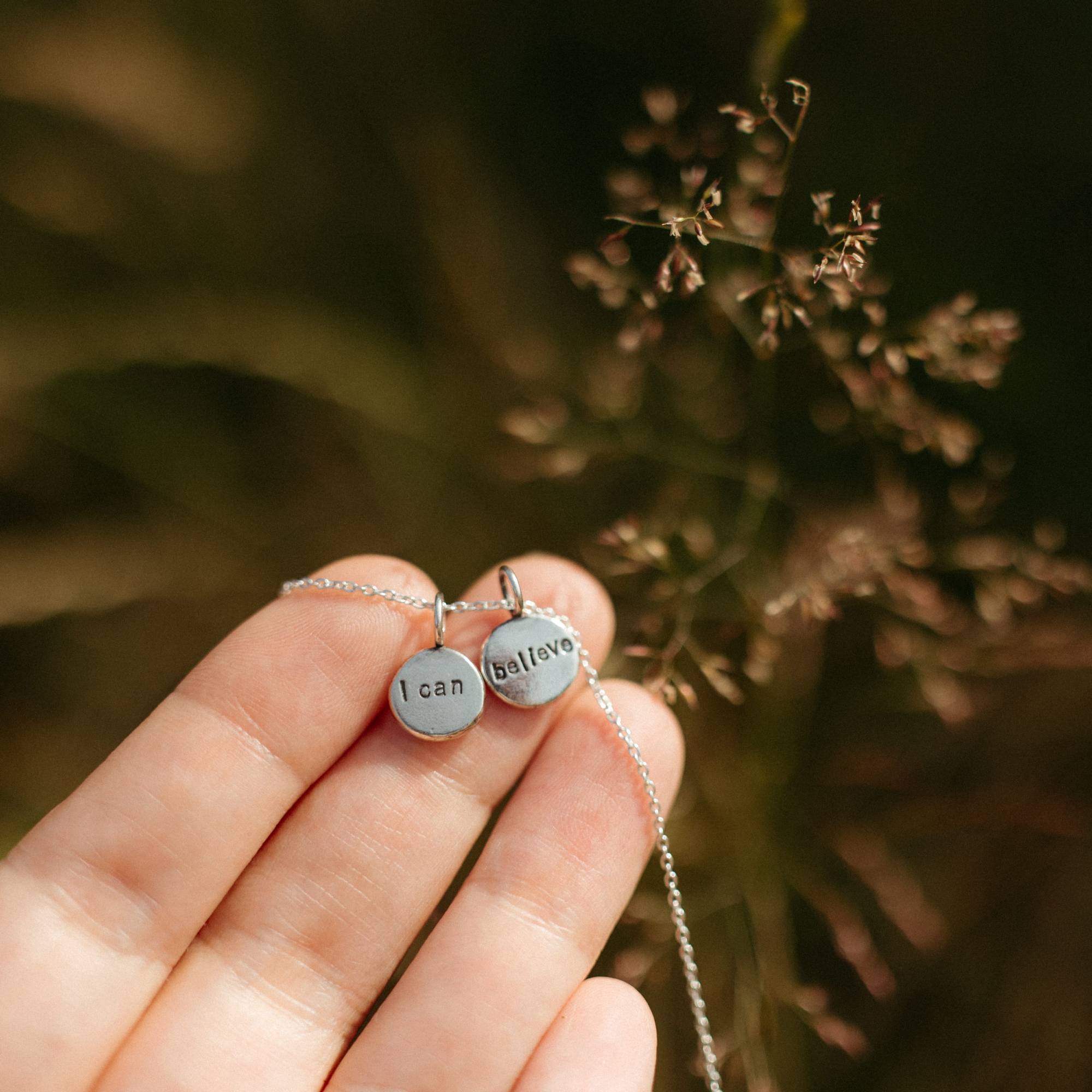 A close-up of a hand holding two small silver round pendant charms on a delicate chain, with one charm engraved 'I can' and the other 'believe'. The background is a soft, out-of-focus natural setting with warm brown and green tones.