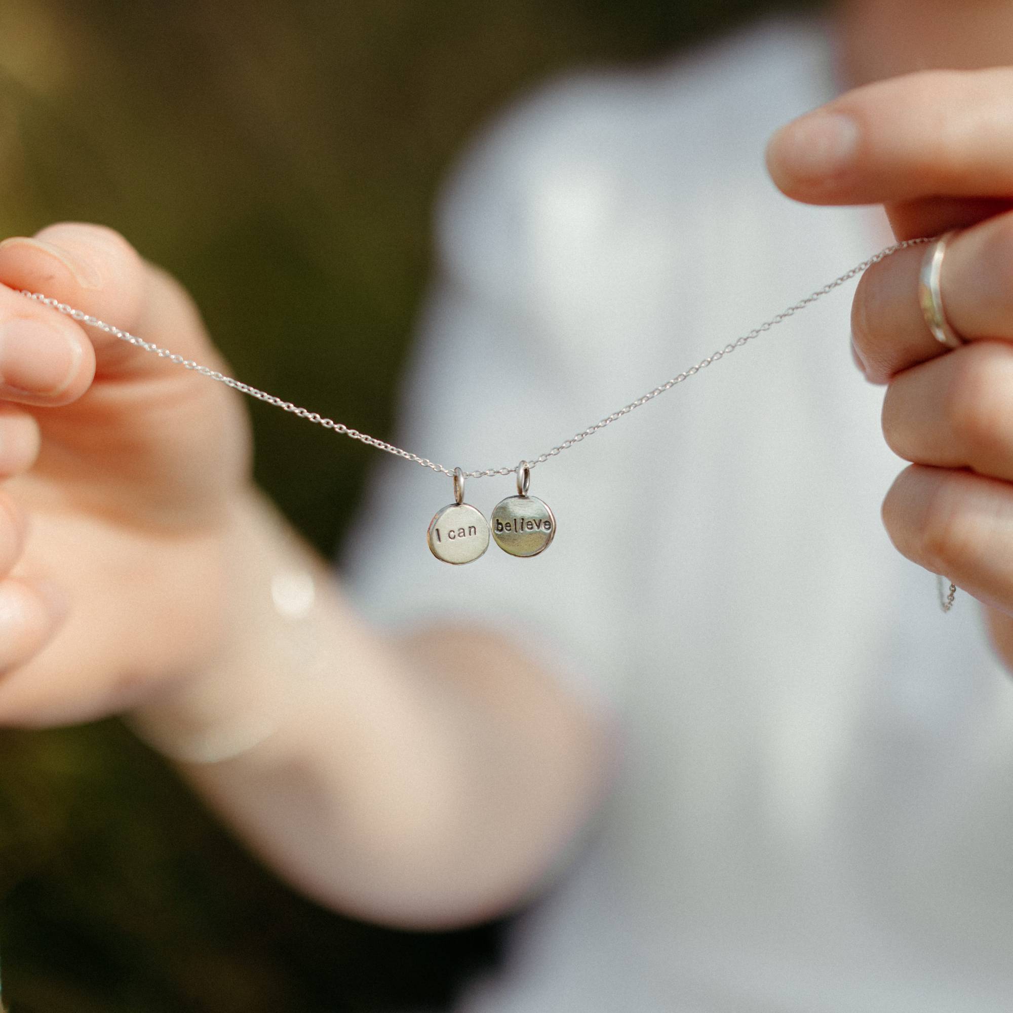 A close-up lifestyle shot of a person holding a delicate silver chain necklace with two small round disc charms engraved with 'I can' and 'believe'. The background is a soft, out-of-focus white and green outdoor setting.
