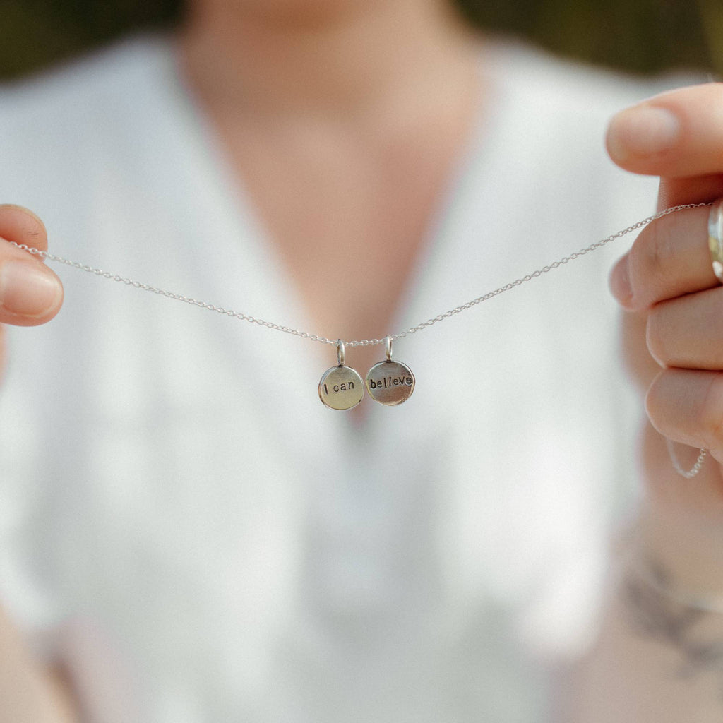 A close-up lifestyle shot of a person holding a delicate silver chain necklace with two small round engraved disc pendants, one engraved with 'I CAN' and the other with 'BELIEVE'. The background is a soft, out-of-focus white shirt.