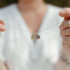 A close-up lifestyle shot of a person holding a delicate silver chain necklace with two small round engraved disc pendants, one engraved with 'I CAN' and the other with 'BELIEVE'. The background is a soft, out-of-focus white shirt.