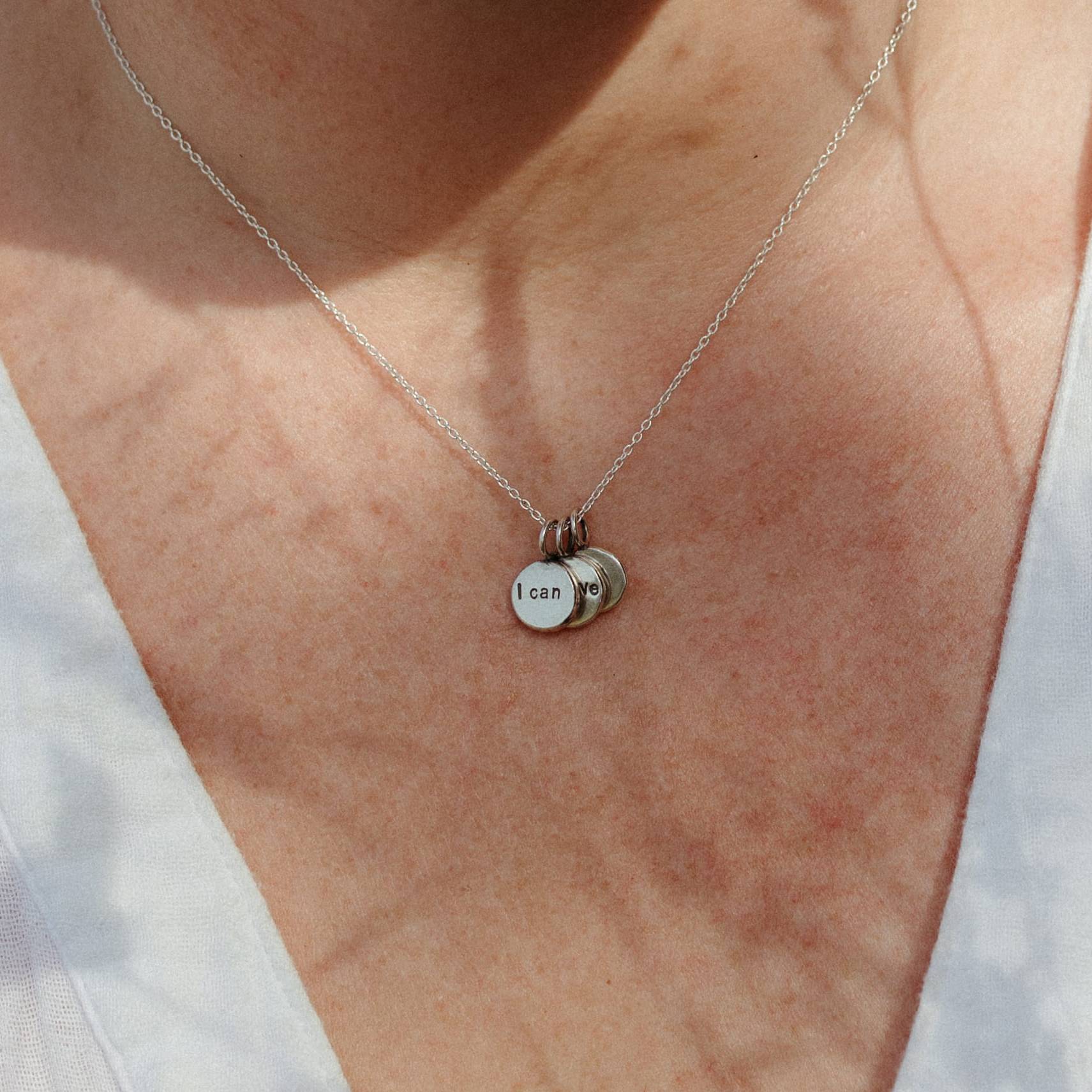 A close-up of a person wearing a delicate silver chain necklace with three small round silver charms, including one stamped with 'I can'.