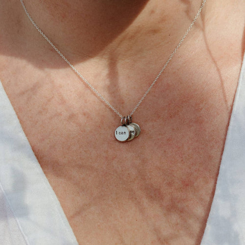 A close-up of a person wearing a delicate silver chain necklace with three small round silver charms, including one stamped with 'I can'.
