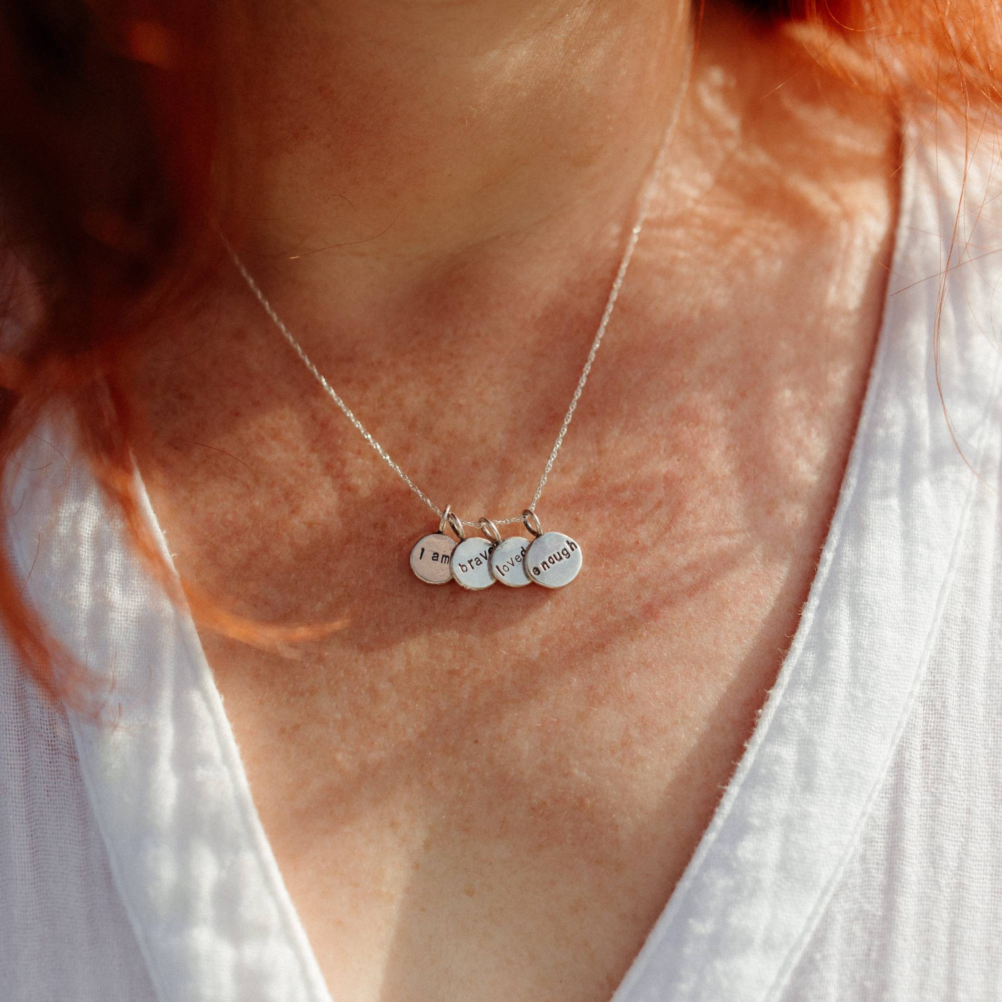 A close-up of a person wearing a delicate silver necklace with four small engraved round charms with the words 'i am', 'brave', 'loved' and 'enough', styled with a white V-neck top.