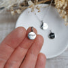 Silver pendant charm with 'align' engraved on the front, held on a hand, with a blurred background of dried flowers and a white dish.