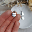 Silver pendant charm with 'believe' hand stamped onbit, held on a hand holding it, with more pendants in the background.