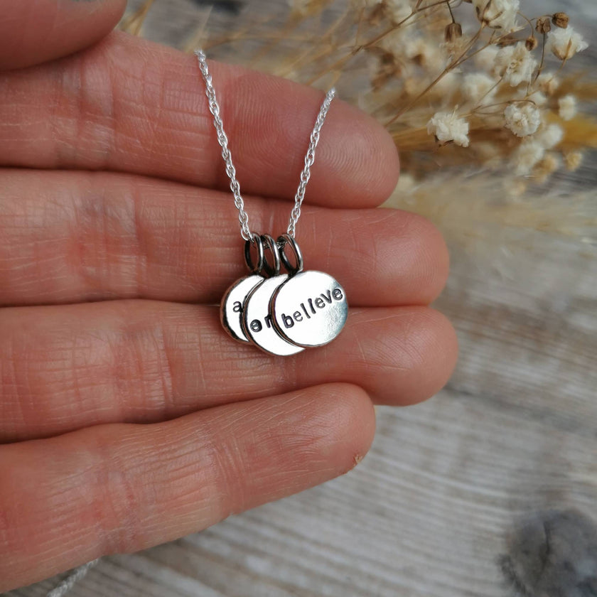 Silver necklace with round charm pendants each with a single word mantra etched into them, held in a hand against a blurred natural background