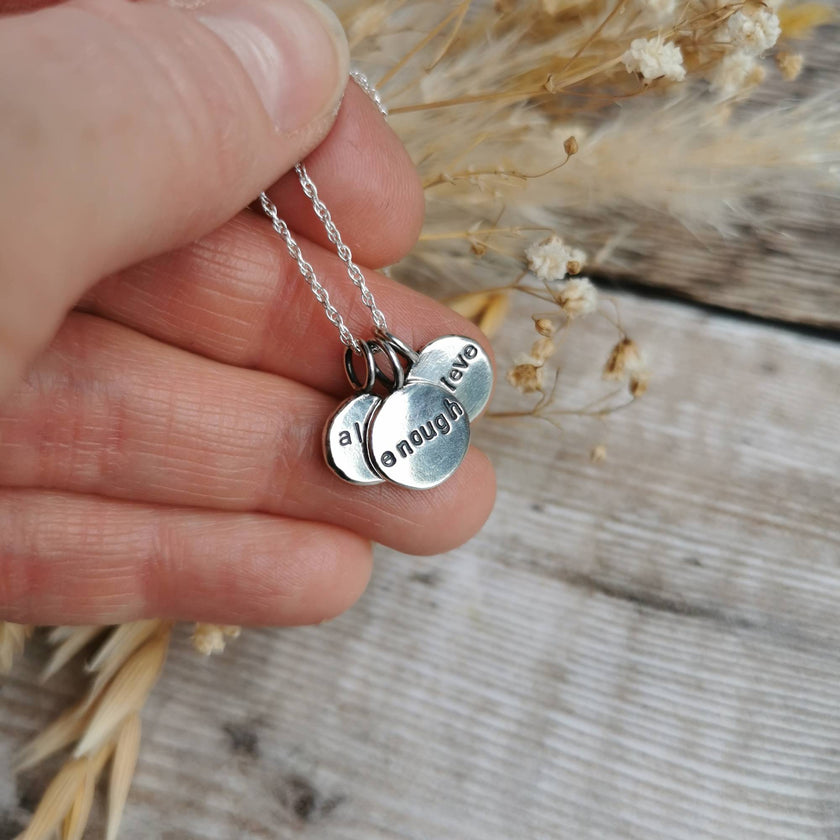 Hand holding a silver necklace with three engraved round charms each with a positive affirmation word stamped into the surface. Held against a natural background
