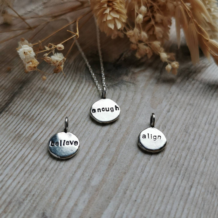 Three silver charm pendants with engraved mantra words on a wooden surface with dried flowers.