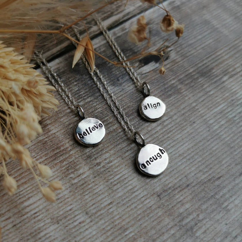 Three silver necklaces with hand stamped charms with positive affirmations of believe, align & enough on a wooden surface with dried grasses