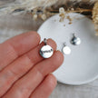 Silver pendant  charm with 'enough' engraved onto the front, held by a hand, with a blurred background of dried flowers and another pendant on a white dish.