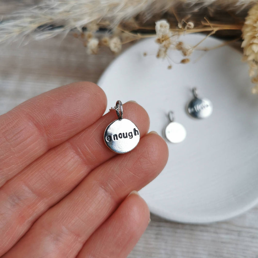Silver pendant  charm with 'enough' engraved onto the front, held by a hand, with a blurred background of dried flowers and another pendant on a white dish.